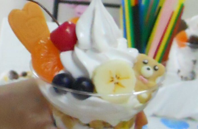 Close-up of a colorful parfait with mix-ins like a bear cookie, banana slices, blueberries, orange, cherry, and other toppings at Sample Riki.